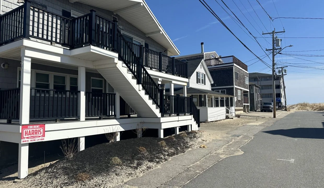 A quiet street lined with two-story residential buildings featuring balconies, with a real estate sign indicating a property for sale.