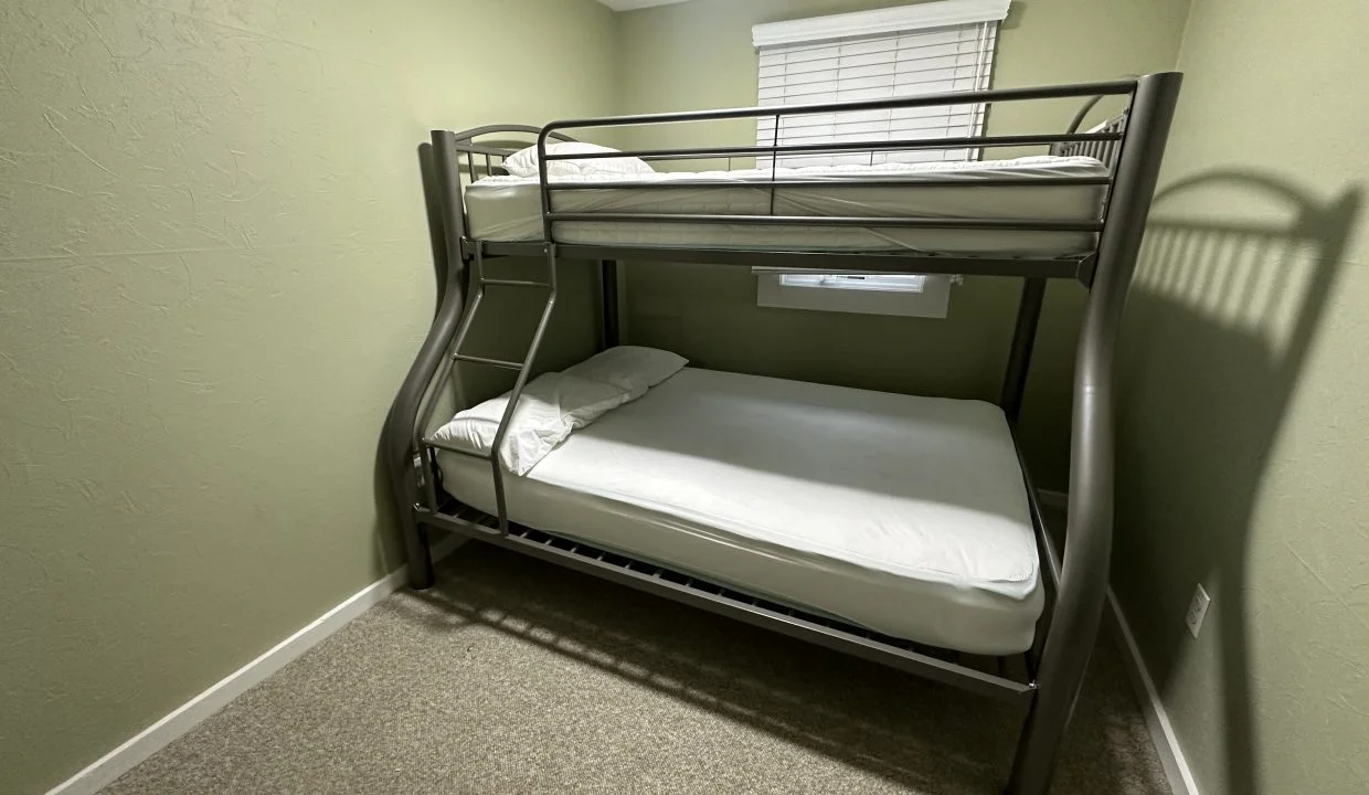 A minimalist room with a metal bunk bed featuring a lower double bed and an upper single bed, positioned against a green wall and under a window.