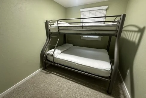 A minimalist room with a metal bunk bed featuring a lower double bed and an upper single bed, positioned against a green wall and under a window.