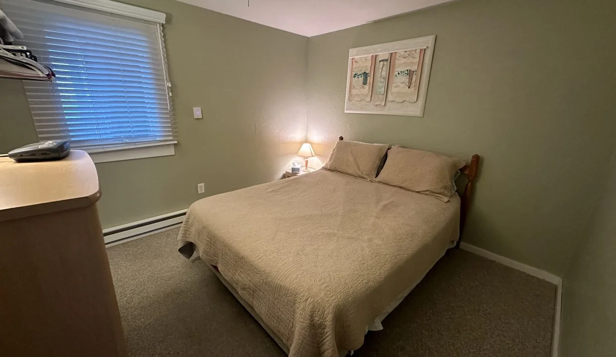 A modestly furnished bedroom with a double bed, beige comforter, two wall art pieces, a small lamp lit, and a window with blinds.