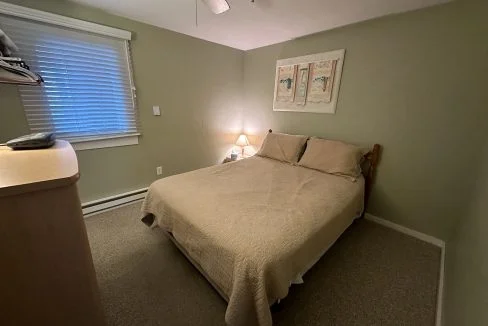 A modestly furnished bedroom with a double bed, beige comforter, two wall art pieces, a small lamp lit, and a window with blinds.
