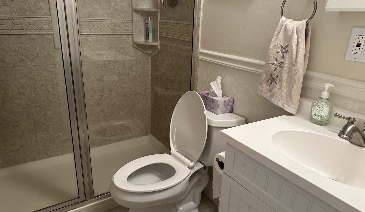 A clean bathroom featuring a toilet with an open lid, a shower stall, and a white vanity with a sink. a towel hangs on the rack and toiletries are on the counter.