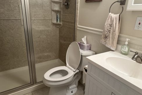 A clean bathroom featuring a toilet with an open lid, a shower stall, and a white vanity with a sink. a towel hangs on the rack and toiletries are on the counter.