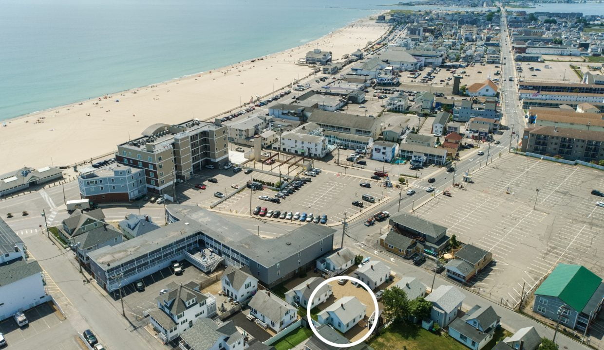 Aerial view of a coastal town with a long sandy beach, numerous buildings, parking lots, and residential houses, one of which is circled. The ocean lies in the background.