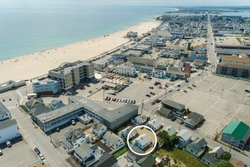 Aerial view of a coastal town with a long sandy beach, numerous buildings, parking lots, and residential houses, one of which is circled. The ocean lies in the background.