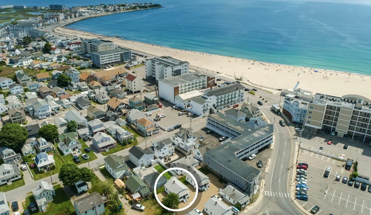 Aerial view of a coastal town with numerous buildings, a sandy beach, and the ocean. A white circle highlights a specific building within the town.