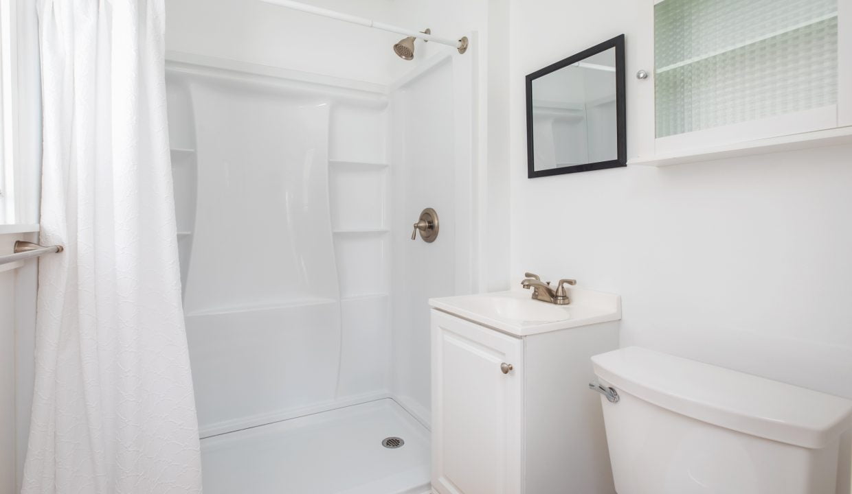 Small, clean bathroom with a shower, white curtain, white sink with silver faucet, white toilet, and mirrored cabinet on the wall.
