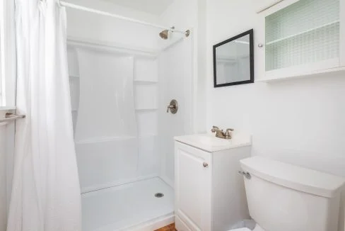 Small, clean bathroom with a shower, white curtain, white sink with silver faucet, white toilet, and mirrored cabinet on the wall.