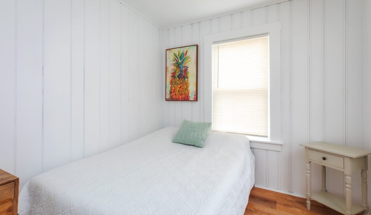 A small bedroom with white paneled walls, a bed covered with a white quilt, one green pillow, a colorful painting, a window with closed blinds, and a wooden nightstand.