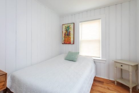 A small bedroom with white paneled walls, a bed covered with a white quilt, one green pillow, a colorful painting, a window with closed blinds, and a wooden nightstand.