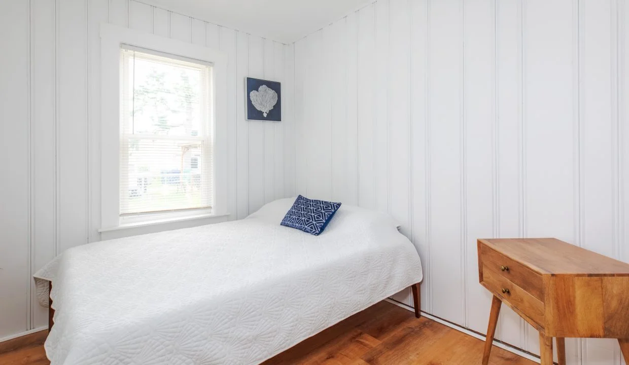 A small bedroom with white paneled walls features a bed covered with a white quilt, a blue pillow, a wooden side table, and a window with blinds. There is artwork on the wall above the bed.