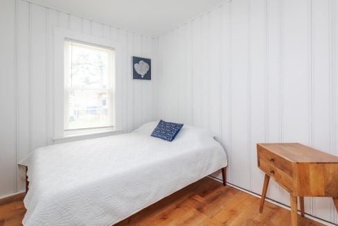 A small bedroom with white paneled walls features a bed covered with a white quilt, a blue pillow, a wooden side table, and a window with blinds. There is artwork on the wall above the bed.