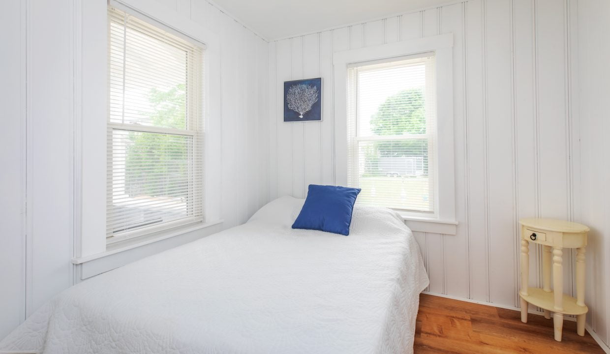 A small bedroom with white wood-paneled walls, two windows with blinds, a bed with a white quilt and a blue pillow, a nautical-themed wall decoration, and a small round yellow nightstand.