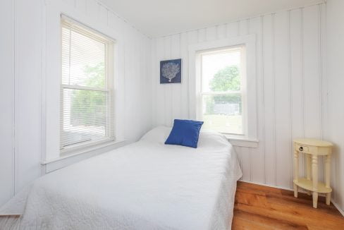 A small bedroom with white wood-paneled walls, two windows with blinds, a bed with a white quilt and a blue pillow, a nautical-themed wall decoration, and a small round yellow nightstand.