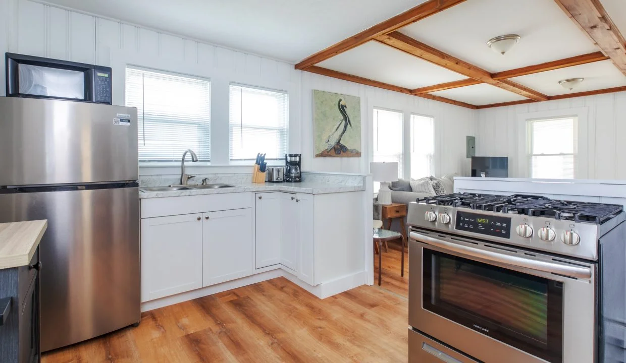 A modern kitchen with stainless steel appliances, including a refrigerator and gas stove, white cabinets, a marble countertop, wooden floor, and a small dining area in the background.