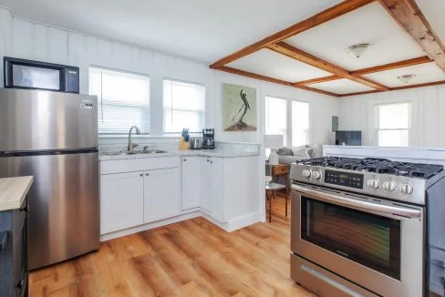 A modern kitchen with stainless steel appliances, including a refrigerator and gas stove, white cabinets, a marble countertop, wooden floor, and a small dining area in the background.