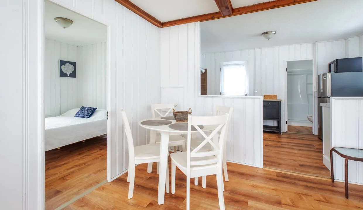 A white dining table with four chairs is set in a small open-plan space with light wooden floors. A bedroom and a kitchen area with modern appliances can be seen in the background.