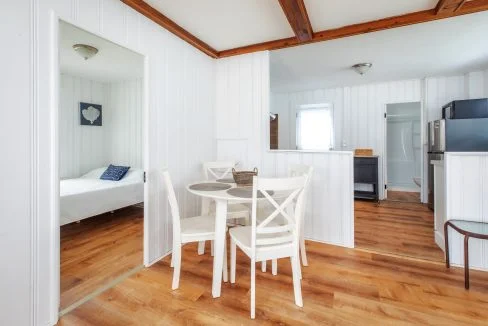 A white dining table with four chairs is set in a small open-plan space with light wooden floors. A bedroom and a kitchen area with modern appliances can be seen in the background.