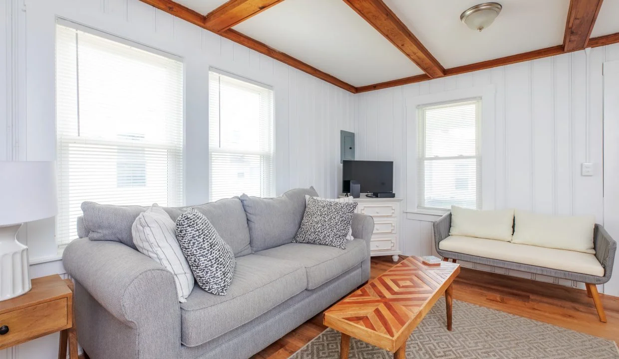 A living room with a gray sofa, patterned cushions, wooden ceiling beams, a small white sofa, a wooden coffee table, and a TV on a stand against a white-painted wall with three windows.