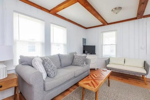 A living room with a gray sofa, patterned cushions, wooden ceiling beams, a small white sofa, a wooden coffee table, and a TV on a stand against a white-painted wall with three windows.