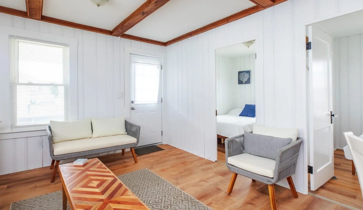 A bright living room with wooden floors, a zigzag-patterned coffee table, a rug, two chairs, a small sofa, and an open door leading to a bedroom. Light streams in through a window and door.