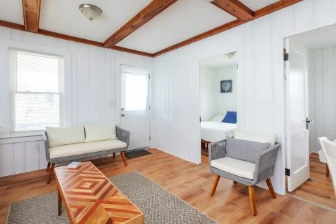 A bright living room with wooden floors, a zigzag-patterned coffee table, a rug, two chairs, a small sofa, and an open door leading to a bedroom. Light streams in through a window and door.