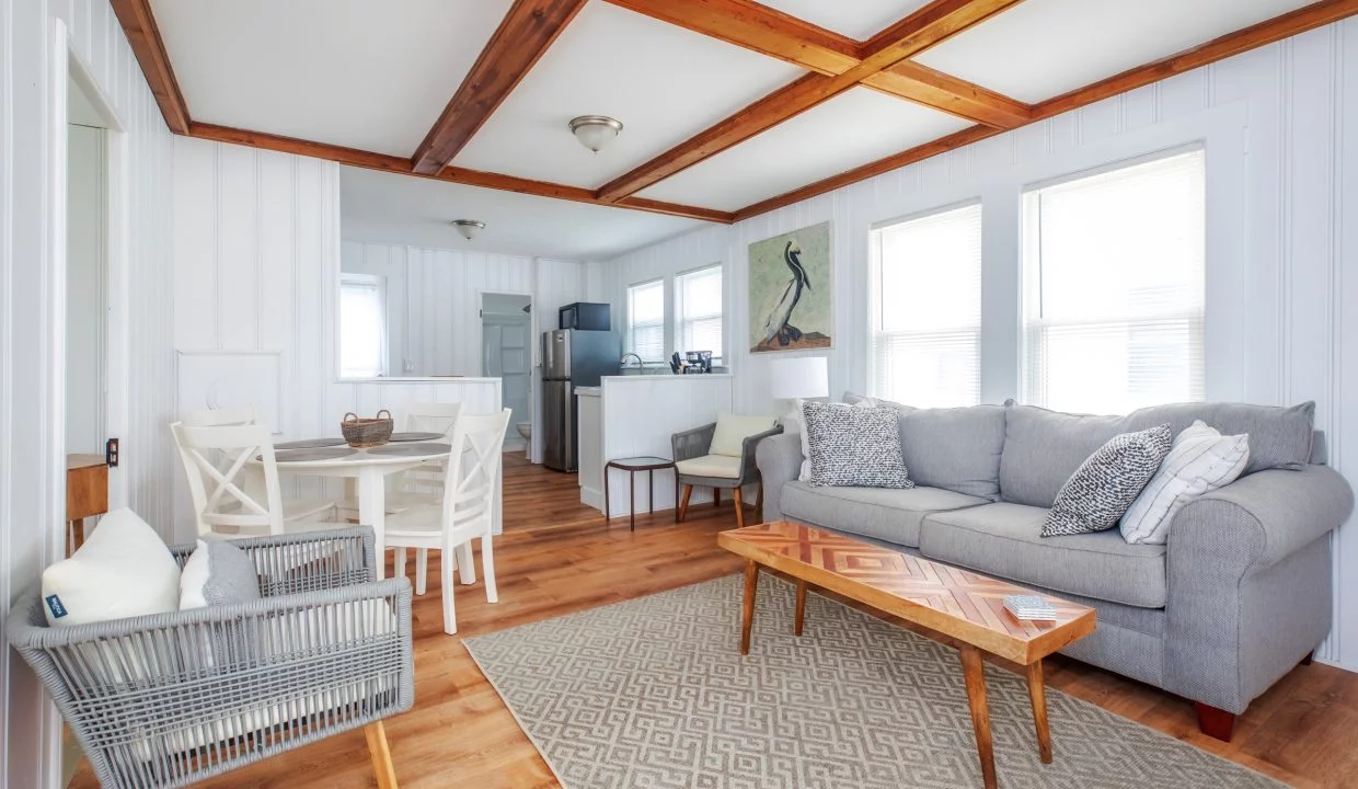 A cozy living room with a gray couch, wooden coffee table, wicker chair, and a dining area. The room features white walls, wood ceiling beams, and large windows letting in natural light.