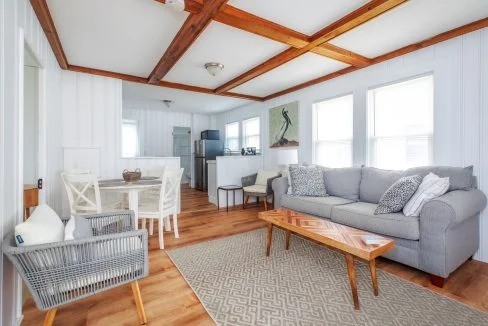 A cozy living room with a gray couch, wooden coffee table, wicker chair, and a dining area. The room features white walls, wood ceiling beams, and large windows letting in natural light.