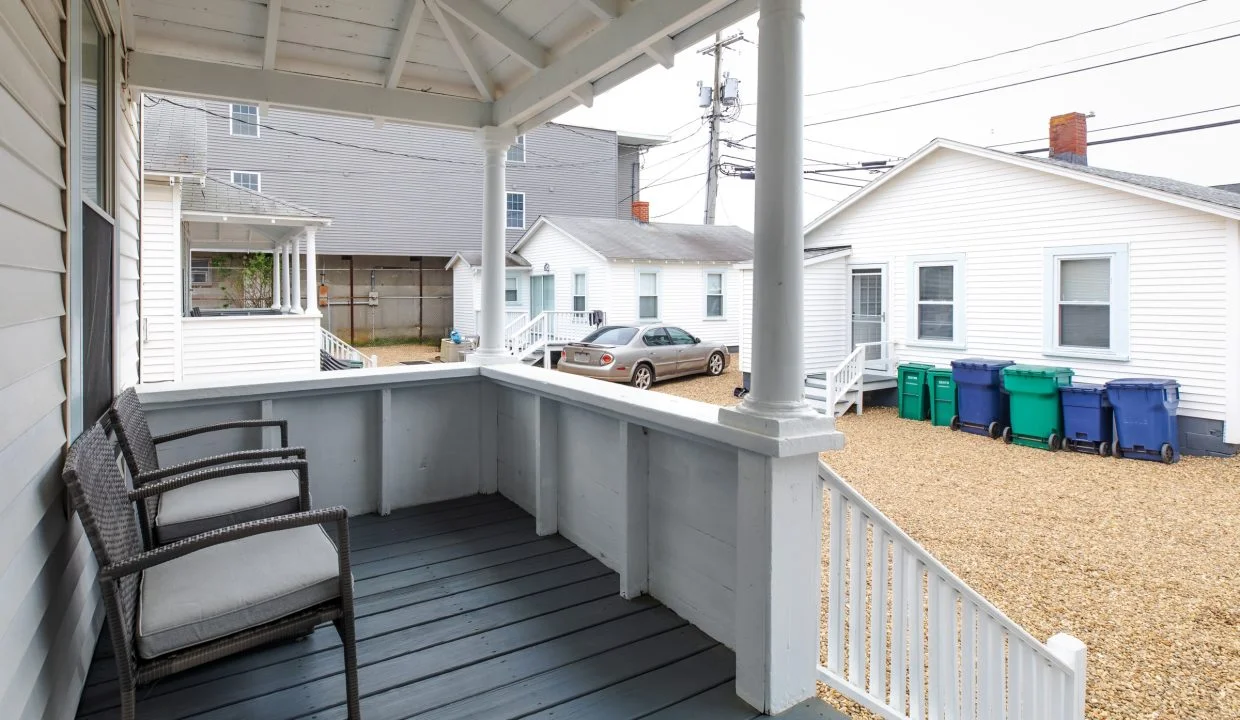 A small porch with two wicker chairs, overlooking a gravel driveway with three recycling bins and parked cars in between white houses.