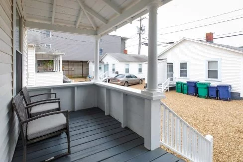 A small porch with two wicker chairs, overlooking a gravel driveway with three recycling bins and parked cars in between white houses.