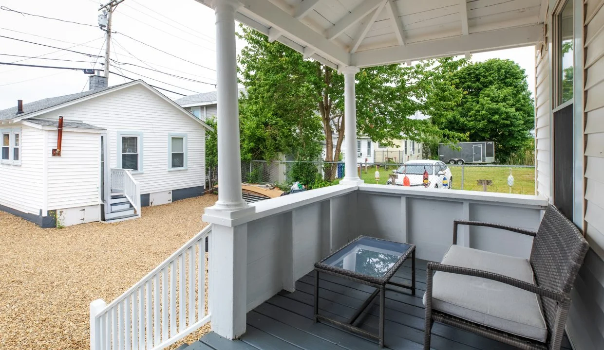 A porch with a wicker chair and glass-top table overlooks a gravel yard with nearby houses and trees.