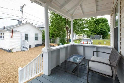 A porch with a wicker chair and glass-top table overlooks a gravel yard with nearby houses and trees.