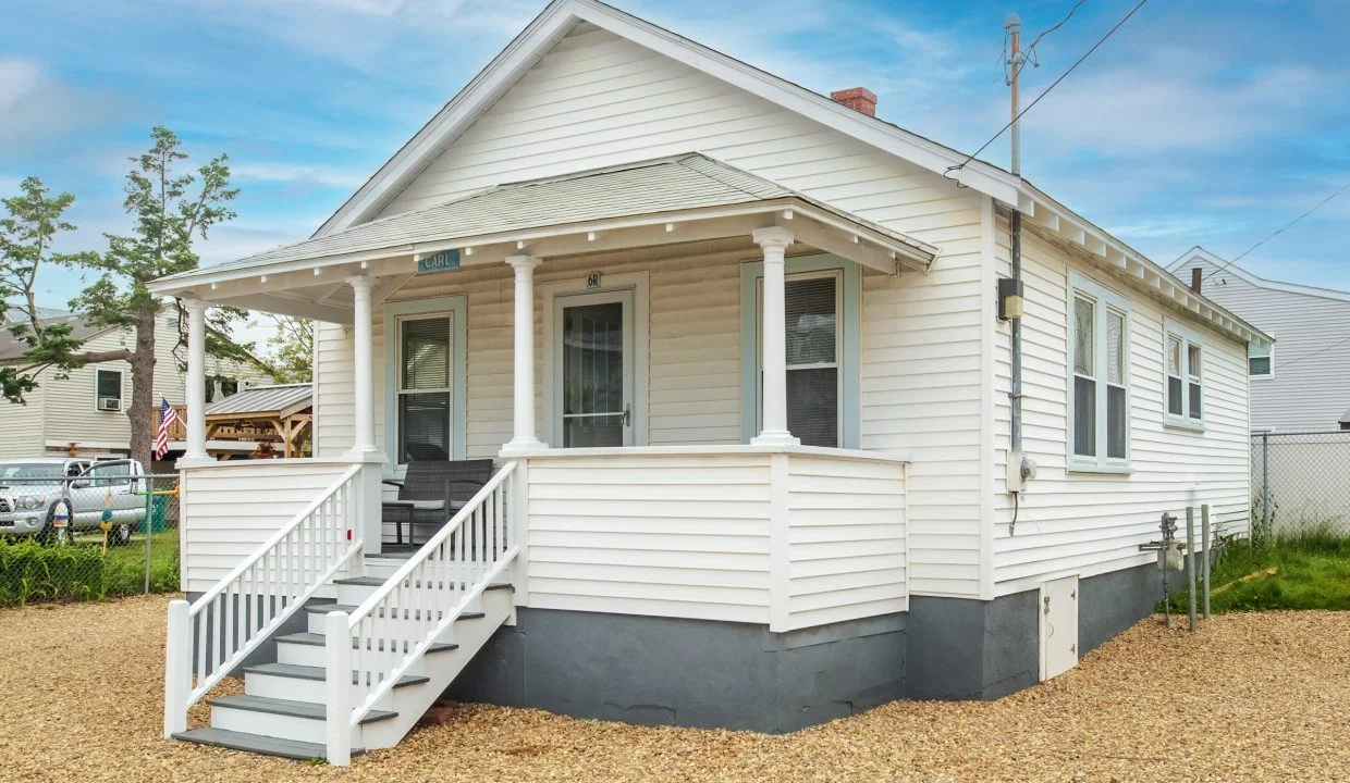 A small, white, single-story house with a porch, stairs leading to the entrance, and a gravel yard.