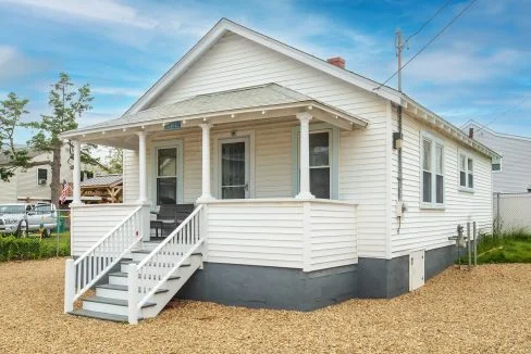 A small, white, single-story house with a porch, stairs leading to the entrance, and a gravel yard.