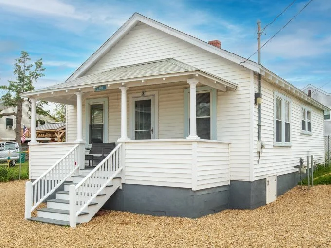 A small, white, single-story house with a porch, stairs leading to the entrance, and a gravel yard.