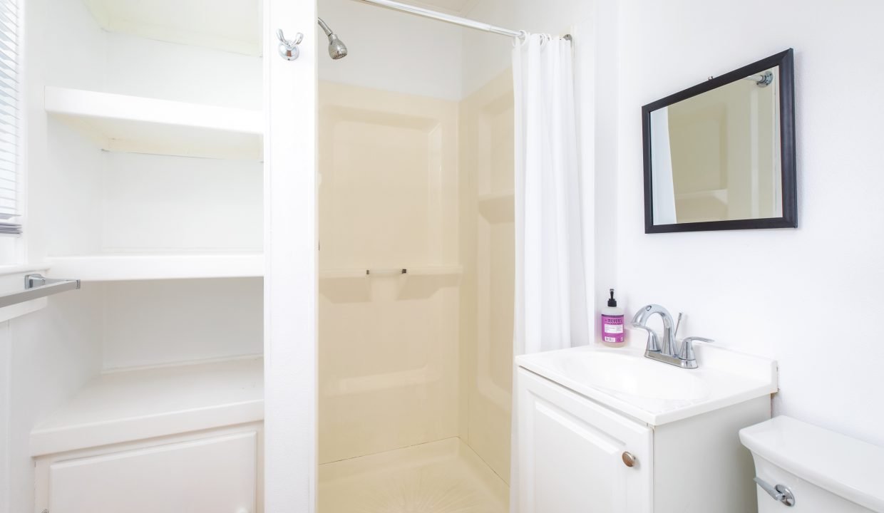 A small bathroom features a shower with a curtain, a vanity with a sink and mirror, white shelves, and a toilet. Sanitizer bottle is on the sink. Walls and furniture are white.