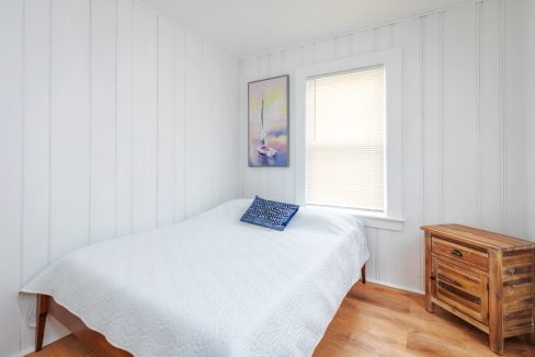 A small bedroom with white paneled walls, a wooden floor, a single wooden nightstand, and a bed covered with a white quilt and a blue pillow. A picture and a window with blinds are on the wall.