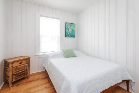 A small bedroom features a bed with a white cover and a green pillow, a wooden nightstand, a window with blinds, and a painting on a white paneled wall.