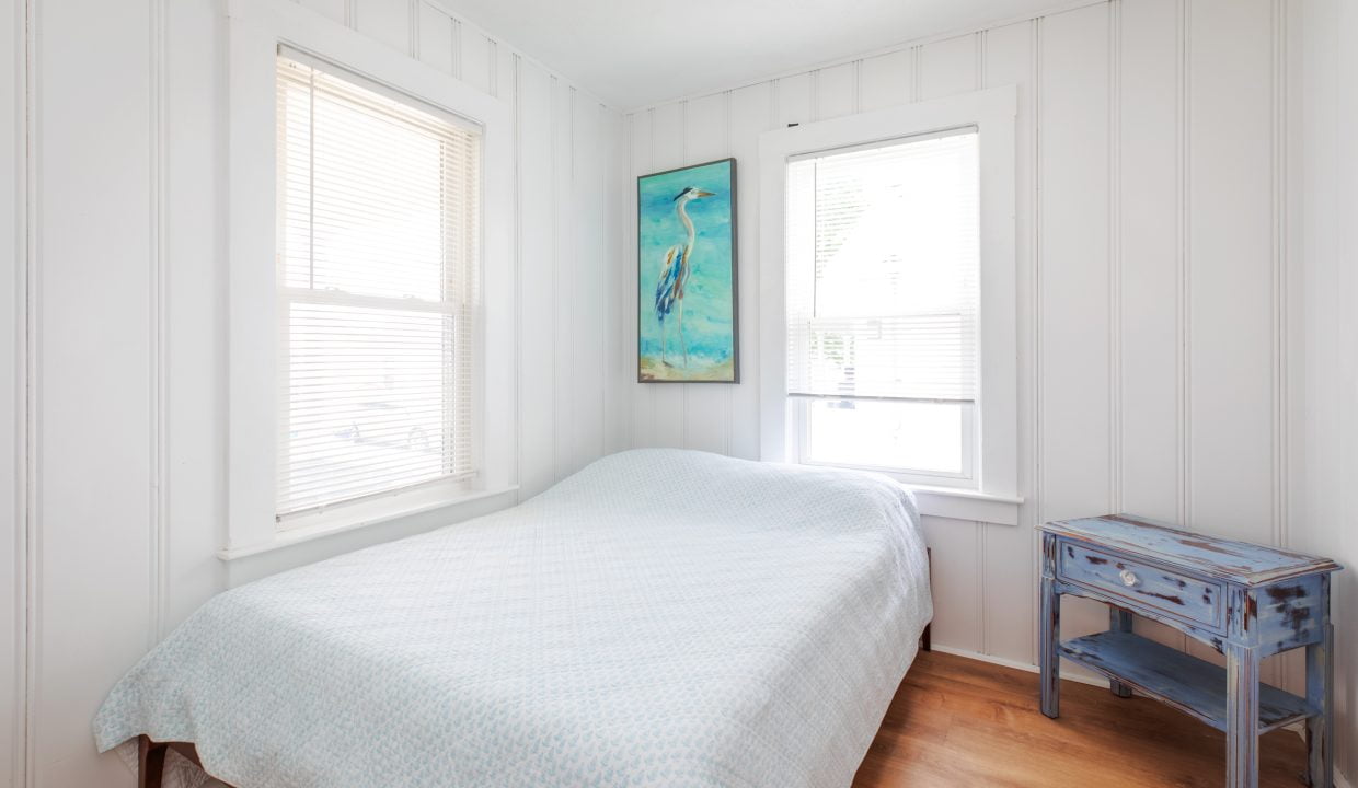 A small bedroom with white walls, a single bed covered with a light blue bedspread, a blue nightstand, and a painting of a heron on the wall between two windows with blinds.