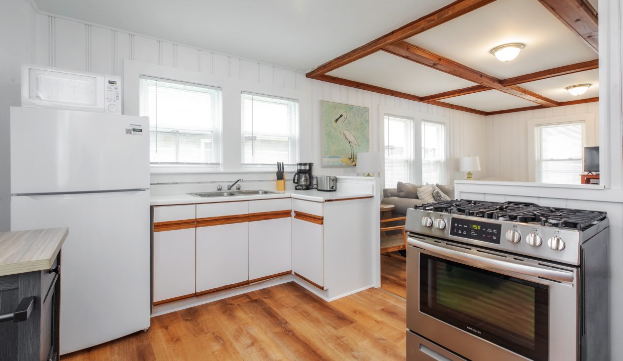 A clean, bright kitchen featuring white cabinets, a refrigerator, a microwave, a stove, a coffee maker, and a double sink. Adjacent to the kitchen is a cozy living area with natural light.