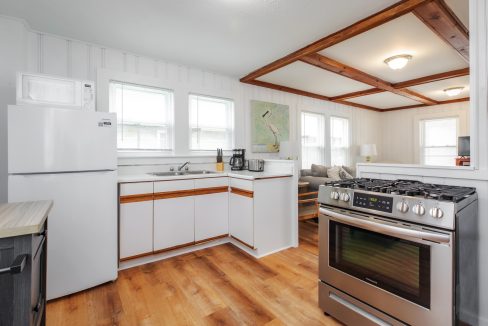 A clean, bright kitchen featuring white cabinets, a refrigerator, a microwave, a stove, a coffee maker, and a double sink. Adjacent to the kitchen is a cozy living area with natural light.