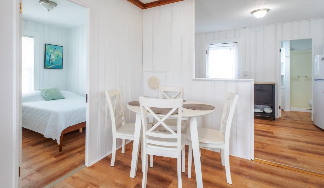 A small dining area with a round table and four white chairs is situated between a bedroom and a kitchen. The room features white walls and wood floors.