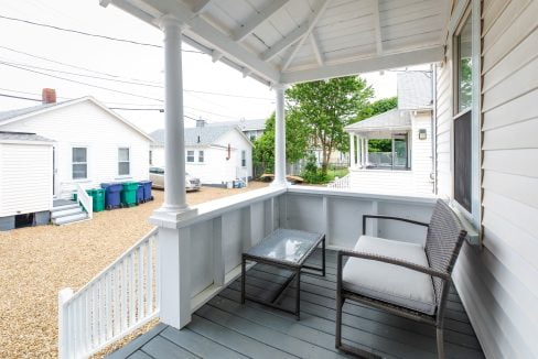 Small covered porch with a wicker chair and glass coffee table, overlooking a gravel yard and neighboring white houses with recycling bins in the background.