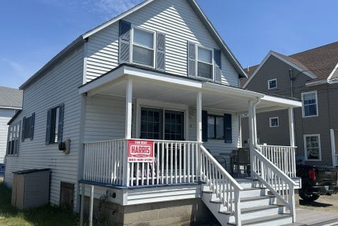 A white two-story house with a large front porch featuring a "For Rent" sign from Harris Realty. The house has blue shutters, a concrete foundation, and a set of stairs leading to the porch.