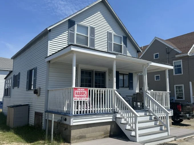 A white two-story house with a large front porch featuring a "For Rent" sign from Harris Realty. The house has blue shutters, a concrete foundation, and a set of stairs leading to the porch.