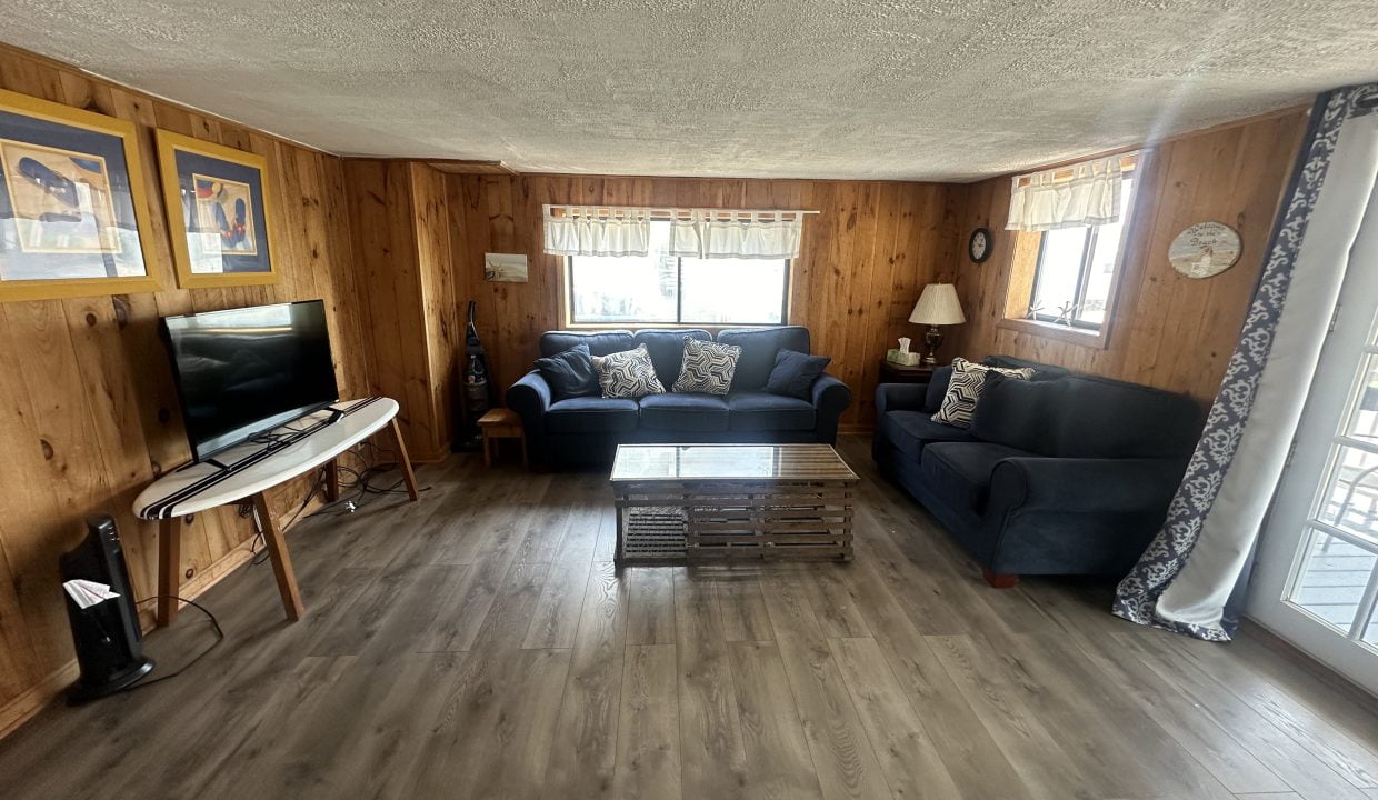 A cozy living room with wood-paneled walls features two blue sofas, a wooden coffee table, a flat-screen TV on a console, and carpeted windows providing natural light.