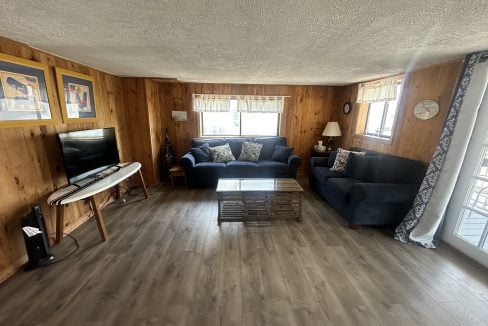 A cozy living room with wood-paneled walls features two blue sofas, a wooden coffee table, a flat-screen TV on a console, and carpeted windows providing natural light.