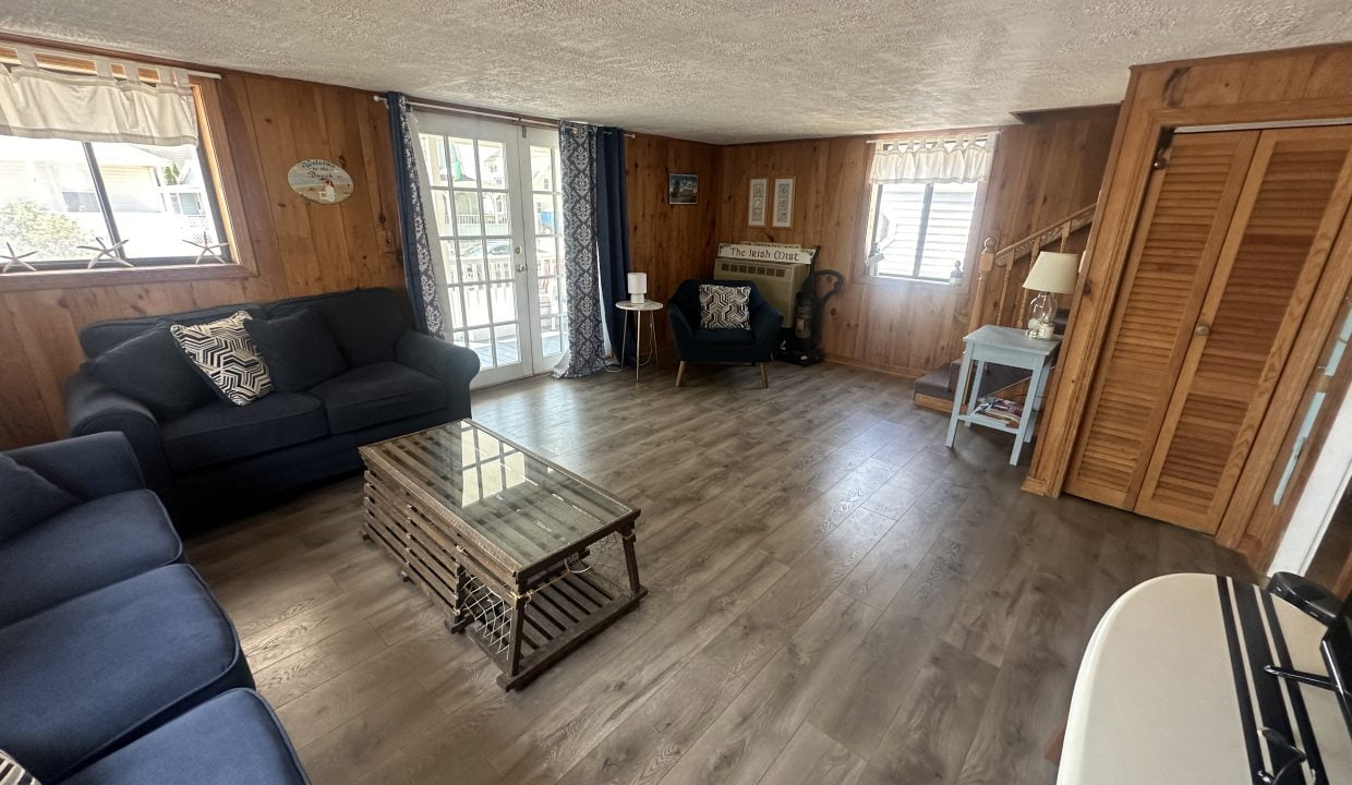 A cozy living room with wooden walls, large windows, a glass coffee table, and navy blue couches arranged around the table. A small desk and chair are situated near the window.