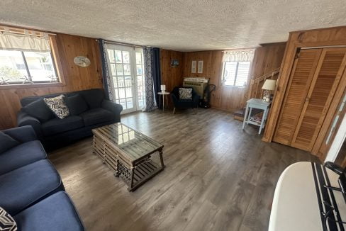 A cozy living room with wooden walls, large windows, a glass coffee table, and navy blue couches arranged around the table. A small desk and chair are situated near the window.