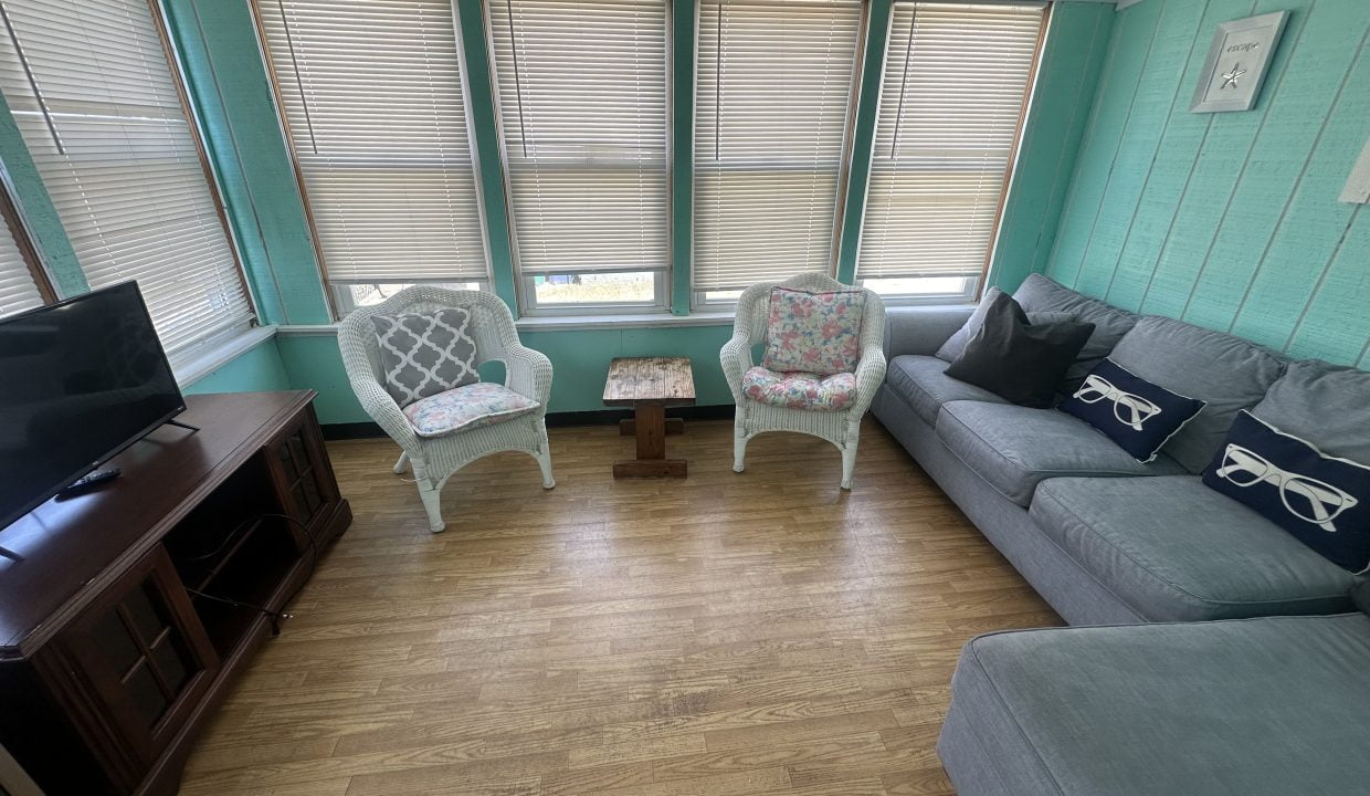 A room with teal walls features a couch, two wicker chairs, a wooden TV stand with a flatscreen TV, and several windows with closed blinds. The floor is wooden.